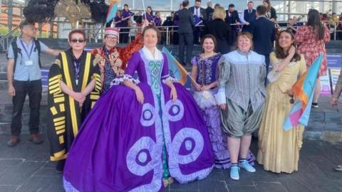 Welsh National Opera performers in bright, colourful dresses and robes. on the steps of the Senedd building.
