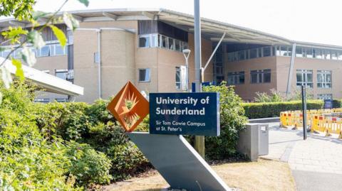 The exterior of a large university building with a blue and orange sign outside reading 'University of Sunderland - Sir Tom Cowie Campus at St. Peter's.