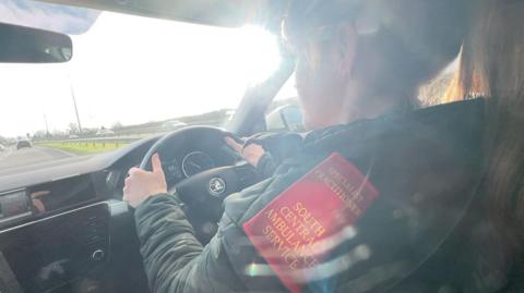 Chrissy Ames is driving her ambulance in the fast lane of a dual carriageway. The sun is shining through the windscreen. Chrissy has both hands on the wheel and her hair tied back in a ponytail. There is gold embroidery on the red epaulette on her shoulder which says specialist practitioner South Central Ambulance Service.