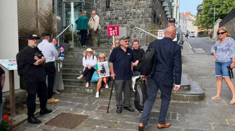 Eleven protestors outside the States with three people holding no to GST placards. A man, Deputy David Dorrity who we see from behind is walking up the stairs to the Royal Court. 