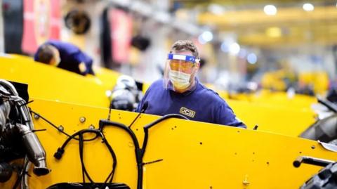 A man in blue overalls and eye protection behind a large yellow machine with more yellow machines behind him