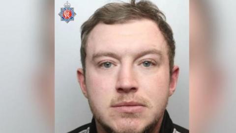 A close-up photo shows the upper part of a person’s head and shoulders against a plain light background. The person has short, light brown hair. In the top-left corner of the image, there is a badge featuring the Greater Manchester Police logo, which includes a red circle with the letters “ER” inside, surrounded by a blue star-shaped design.