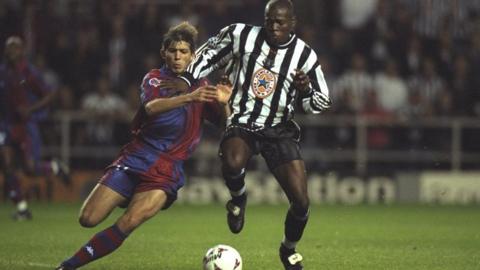 An action shot from the 1997 match between Newcastle and Barcelona. Faustino Asprilla is running with the ball in a long-sleeved black and white Newcastle strip. A Barcelona player is about to slide for the ball in a blue and red strip. Asprilla has his hand on the chest of the Barcelona player.