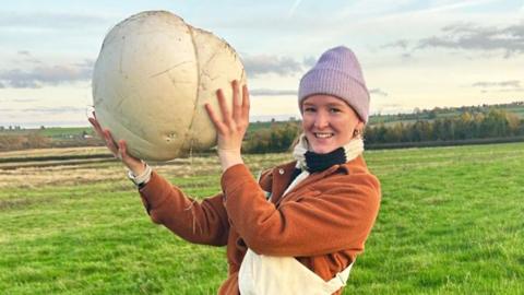 Alissimon Minnitt is wearing a burnt-orange coat and a lavender winter hat. She is standing in a bright green field holding a huge white mushroom, which is three times the size of her head.