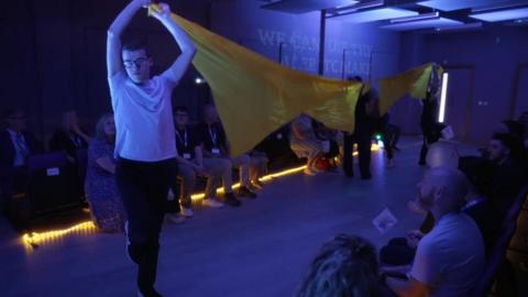 Students hold up a large yellow banner whilst an audience looks on in the new performing arts centre at Prior's Court.