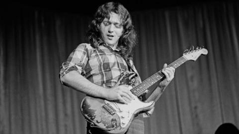 Musician Rory Gallagher in a black and white photograph. He is wearing a checked shirt and holding a guitar with his his head tilted towards the ground. 