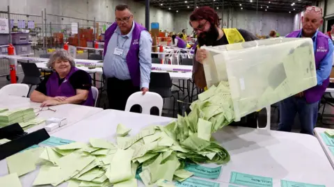 Australian Electoral Commission staff prepare to count ballots during the 2025 federal election at OPC in Brisbane, Australia, May 3, 2025. One worker is dumping a bucket of paper votes on to a table as three more AEC workers look on.