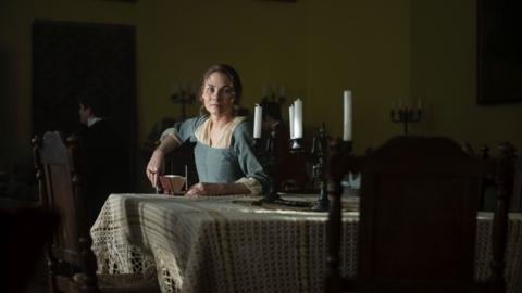 A woman playing Jane Austen sits at a table looking at the camera.