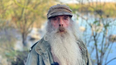 A man with a long white beard and wearing a cap is looking into the camera. He is standing in front of a pond.
