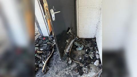 Burnt items are lying on a floor next to a door. They are black and charred. The white wall has black specks on it.