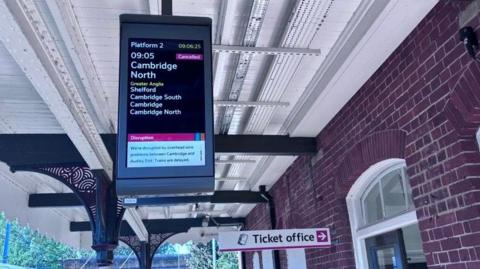 A notice board at a train station is showing a service at 09:05 which has been cancelled. There is a sign which says ticket office which is pointing to a building on the left.