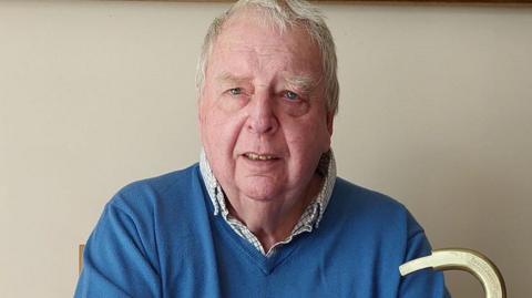 Michael Hamson pictured inside the house. He wears a blue jumper over a white shirt and has short white hair. 