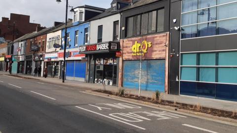 A street view of Holmeside which is a row of shops. Most of the shops have their shutters down and there are a few people walking along the street in front of them.