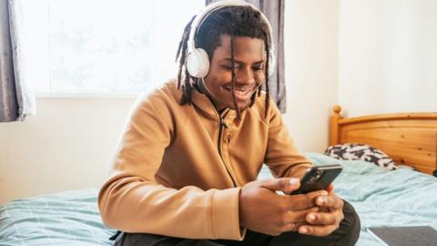 GCSE Biology student on his bed at home wearing big headphones and looking at his phone, listening to a BBC Bitesize revision podcast.