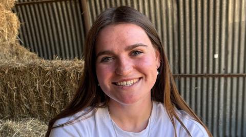Lydia Law looking directly to camera and smiling. She is wearing a white, round-necked t-shirt and has straight brown hair, worn down and reaching over her shoulders. She is seated in a hay barn, with bales of hay see behind her on the left of frame and a corrugated metal wall in the further distance.