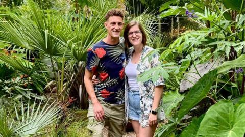 Dave Robinson (left) and Gemma Furby (right) stood in their garden. They are surrounded by large green leaves. Dave is wearing a navy blue t-shirt with red flowers and green leaves on it. He is also wearing khaki green shorts. Gemma is wearing a white vest, blue denim shorts and a white short with green leaves on it.