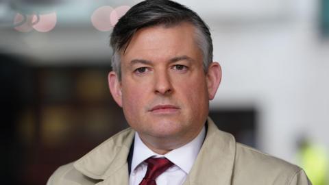 Jonathan Ashworth wearing shirt and tie and a cream coat
