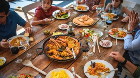 Adults and children sit around a table with various dishes.