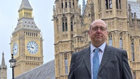 A man with very short grey hair and glasses is dressed in a black suit jacket, white shirt and blue and white tie. He is standing in front of Big Ben and another large building.