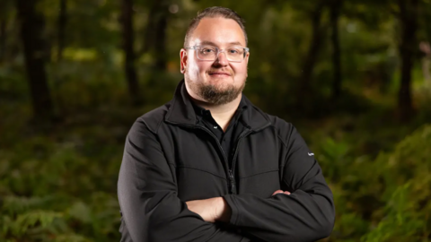 A man in glasses with short dark hair and a beard stands in front of a green foilage background, smiling. He is wearing a black jacket over a black polo shirt, and his arms are folded.