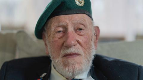 A man looking at the camera. It is a head and shoulders shot of him. He is an older gentleman, he has a grey beard and a green beret on. He has a dark blue suit jacket on, and under that you can see the collars of a white shirt. 