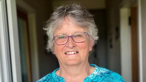 A woman with grey hair wearing glasses wearing an aquamarine top