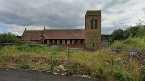 An overgrown area of derelict land behind a temporary wire fence. Grass, weeds and rubble can be seen alongside a stone church with a red roof.