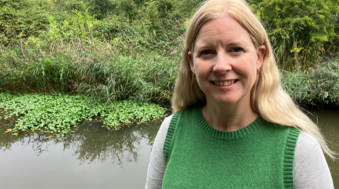 Karen Lewing has long, blonde hair and is wearing a green tank top. She is standing by a canal in Worcester with the invasive plant in the water behind her, which is growing like a green carpet against the banks of the canal