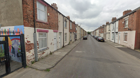 The road has rows of terraced houses on either side. They are red brick and white concrete. To the left, there is a colourful mural on the side of one of the houses.