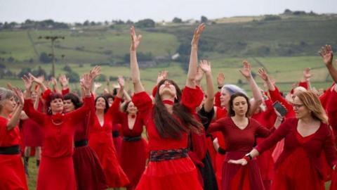 A group of women dressed in red dressed dance on the moors, many with their hands in the air, as part of an event to celebrate Kate Bush