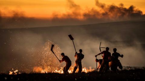 Five firefighters beat a fire on moorland at sunset.
