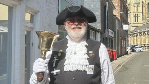 A town crier in ceremonial dress including a black hat and white laced shirt holds a gold bell and smiles at the camera in the street.
