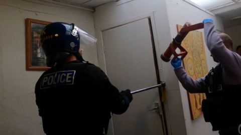 Two police officers can be seen forcing open a door. One has a red battering ram, the other an angled steel tool jammed in the doorframe