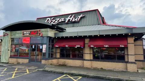 The exterior of a Pizza Hut restaurant with a grey roof displaying a large 'Pizza Hut sign' and maroon window awnings on the windows.
