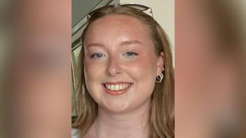 An image of Stephanie Irons, a white woman in her 20s, wearing sunglasses on her head, pushing her hair back and silver hoop earrings. She is smiling.