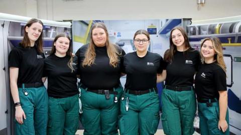 Six university of Worcester paramedic students are standing in a line with their arms around each other. They are all wearing black t-shirts and green trousers