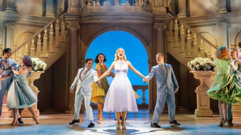 A production image of Helen George on stage, wearing a white dress with her hands being held by cast members as she sings