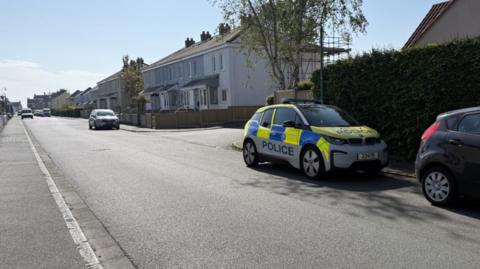 A sunny street, with a number of houses seen on one side. A police car is parked in the foreground.