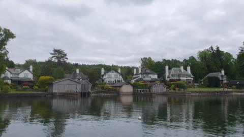 Windermere Villas comprise several large houses on the shores of the lake, some with boathouses. They are pictured from the still, dark water. A mature area of thick trees sits behind the six houses which are detached with a decent gap between each. In the main they are two-storey buildings with additional dormers and have extremely neat gardens dominated by very well-maintained hedges. The wooden, shed-like boathouses, four of which are shown, face out on to the lake and resemble double door garages with pitched roofs.