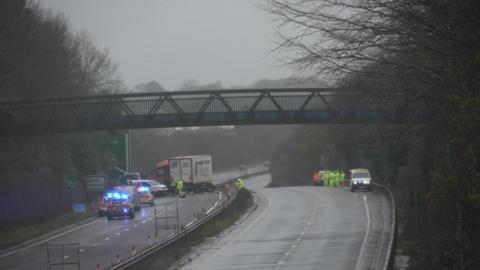 The scene of the collision on the A30. There is a highways maintenance van which has its rear end destroyed. A highways maintenance small lorry is near it and there is a HGV which reads 'Conway Bailey Transport' on the side. There is a stationary police car with flashing lights behind that lorry. Debris covers the carriageway. A road sign reads Bodmin A30 straight on and left turning to Launceston, Bude, Callington and Liskeard.