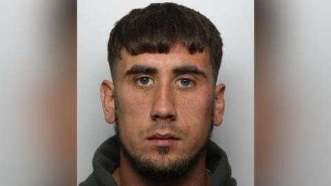 A mugshot of a man who looks straight at the camera. He has brown hair, combed over his forehead and facial hair. He is wearing a jumper.
