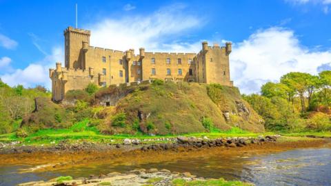 The castle is bathed in sunshine and sits on a rocky outcrop above a loch.