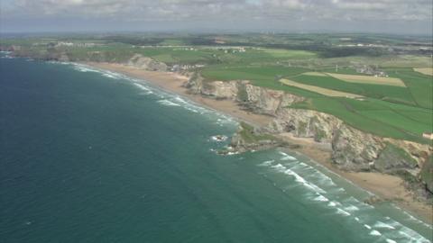 A drone shot of Watergate Bay in Cornwall. It shows the sea as well as green fields in the background.