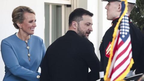 Ukrainian President Volodymyr Zelensky, dressed in black, leaves the White House as a woman shakes his hand and a member of the military stands nearby with a US flag in the foreground of the image