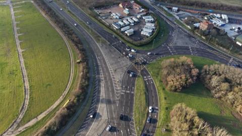 An aerial shot of a roundabout which is in the bottom right of the image and has trees planted on it. A dual carriageway is on the left of the image and merges on to the roundabout.