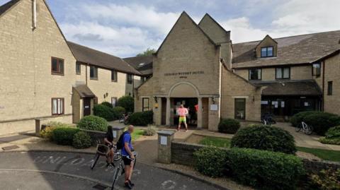 Outside the Witney Four Pillars Hotel on a cloudy day. A couple of people are seen on bicycles in front of the building. Another man is standing at the entrance.