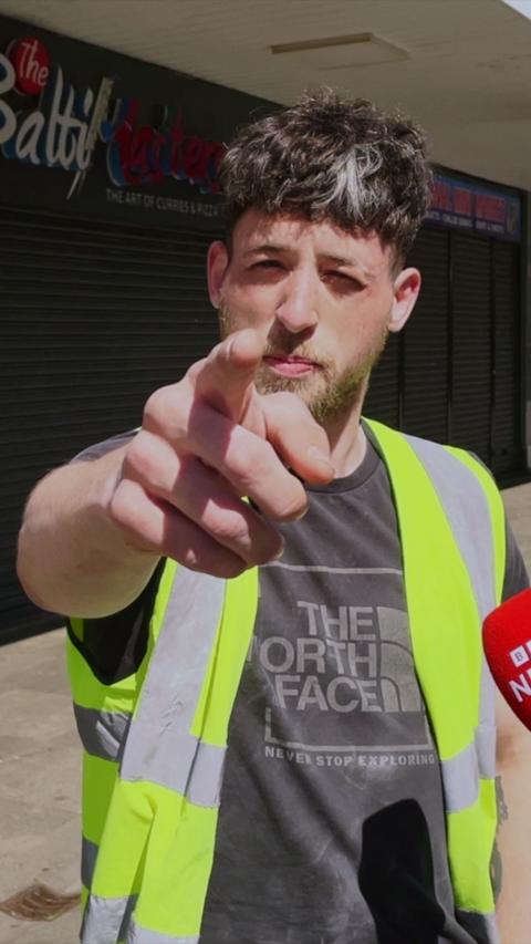 A person wearing a high‑visibility vest points directly towards the camera while standing on a sunlit pavement outside closed shopfronts