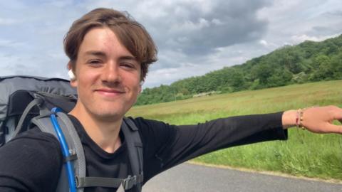 A young man skateboards along an open road lined by long grass and trees. The skateboard is not visible but he has his arms outstretched to balance himself