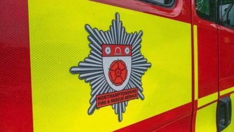 A close up of the side of a red and yellow Northamptonshire Fire and Rescue Service vehicle, with its crest on the side. The crest has a red Tudor rose in the middle.