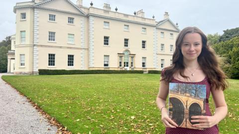 A woman looks towards the camera, stood on a patch of grass in front of a large house. She is holding a book with a photograph on the front. The woman has brunette hair and she is wearing a burgundy top.